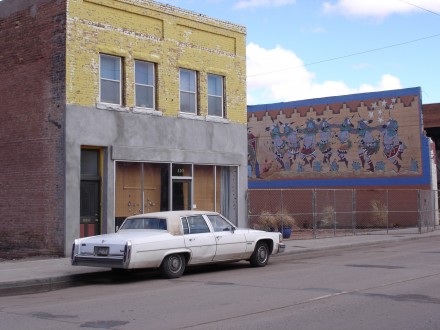 A white Cadillac sits idle, outside a yellow brick building, facing a mural of Indian dancers in Winslow, Ariz. ©Joshua Brockman 2005. All Rights Reserved.